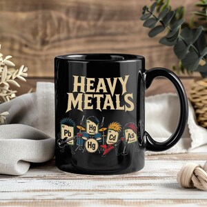 Puede incluir: Una taza de cerámica negra con el texto HEAVY METALS en una fuente beige negrita. Debajo del texto, un gráfico representa cuatro símbolos de elementos químicos antropomórficos como una banda de rock, incluyendo Pb, Hg, Cd y As, tocando instrumentos como guitarras y batería.