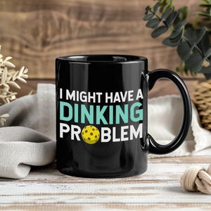 May include: Black ceramic mug with the text "I MIGHT HAVE A DINKING PROBLEM" in white and teal. A yellow pickleball is featured in the design. The mug has a black handle and is set against a wooden background.