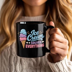 May include: A black ceramic mug featuring a graphic of a three-scoop ice cream cone in blue, purple, and pink. The text reads Ice Cream SOLVES EVERYTHING in blue and pink lettering, accented with small white hearts and stars on the mug surface.