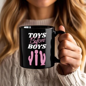 Può includere: Tazza in ceramica nera con la scritta "TOYS Before BOYS" in bianco e rosa. Sotto il testo ci sono tre illustrazioni di giocattoli rosa. La tazza ha un manico nero ed è tenuta da una persona.