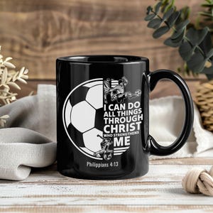 May include: Black ceramic coffee mug with a soccer ball graphic and the text "I CAN DO ALL THINGS THROUGH CHRIST WHO STRENGTHENS ME" and "Philippians 4:13". The mug has a curved handle.