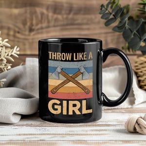 May include: Black ceramic mug with the text "THROW LIKE A GIRL" in a retro design. The design includes crossed axes and horizontal stripes in shades of blue, orange, and red. The mug has a large handle.