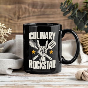 以下が含まれることがあります： 白い大文字で「CULINARY ROCKSTAR」と書かれた黒いセラミックマグカップ。中央にはロックンロールのハンドサインをする手のイラストが描かれ、木製のスプーンとスパチュラが交差しており、両脇に2つの黄色い星が配置されています。