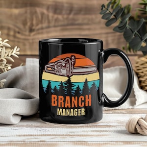 Pode incluir: Caneca de café preta com um gráfico de motosserra em um design de pôr do sol. O texto "BRANCH MANAGER" é exibido em laranja e branco. A caneca tem uma alça preta e está em uma superfície de madeira.
