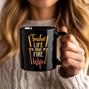 Può includere: Tazza in ceramica nera con la scritta "Teacher Life and Fire Wife" in giallo, arancione e rosa. Il design include immagini stilizzate di una matita e un'ascia. La tazza ha un manico nero.