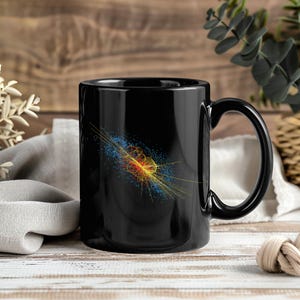 May include: A black ceramic mug featuring a colorful graphic of a particle physics collision event. The design displays a central cluster of yellow and orange circular lines surrounded by a spray of blue and yellow particles on a black background.