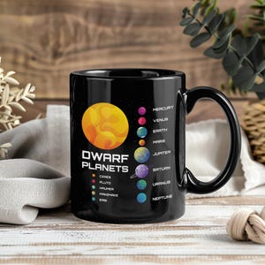 Peut inclure: Mug en céramique noire avec une illustration colorée des planètes naines et des planètes de notre système solaire. La tasse comprend le texte "DWARF PLANETS" et les noms des planètes.