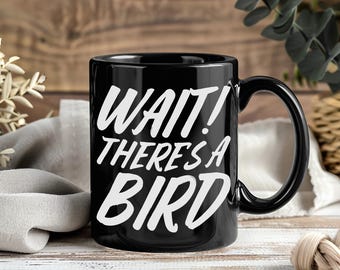 May include: Black ceramic coffee mug with the text "WAIT! THERE'S A BIRD" in white. The mug has a large handle and is set against a wooden background with greenery.