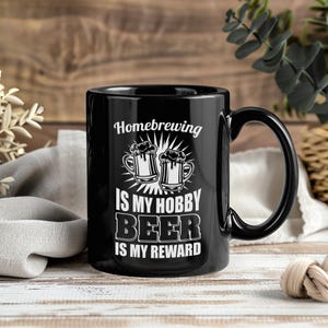 May include: Black ceramic mug with the text "Homebrewing is my hobby, beer is my reward" in white. The mug features an illustration of two beer mugs. The mug has a black handle and is set against a wooden background.