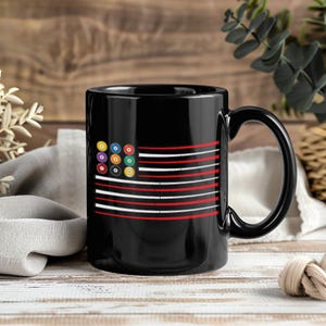 May include: Black coffee mug featuring a design that resembles the American flag, with billiard balls replacing the stars. The stripes alternate between red and white. The mug has a large handle and is set against a wooden background.