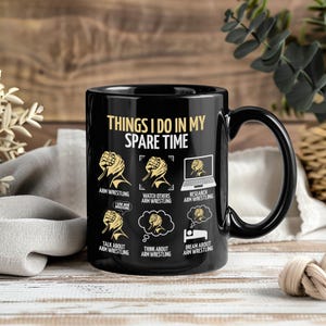 May include: Black ceramic mug with a curved handle, featuring the text "THINGS I DO IN MY SPARE TIME" in gold lettering. The mug has illustrations of arm wrestling and related activities. A humorous gift for arm wrestling enthusiasts.