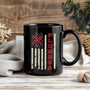 Könnte beinhalten: Schwarze Keramik-Tasse mit einem verwitterten amerikanischen Flaggen-Design, einem roten Schraubenschlüssel und dem Wort "PLUMBER" in Rot. Die Tasse hat einen großen, gebogenen Griff.