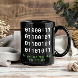 Peut inclure: Mug noir avec code binaire blanc et le texte "If you can read this, you are too." La tasse a une grande anse et est posée sur un fond en bois.