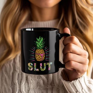 May include: Black coffee mug featuring a cartoon pineapple wearing a pink bikini. The pineapple is surrounded by pink and blue rays, with the word "SLUT" in colorful floral letters below. The mug has a black handle.