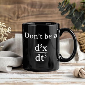 May include: A black ceramic mug featuring the white printed text "Don't be a" above a mathematical derivative symbol representing the third derivative of x with respect to time, d³x/dt³, which is commonly known as a jerk in physics.