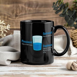 May include: Black ceramic mug with a handle, featuring a graphic of a glass half full of blue liquid. Text on the mug reads "Pessimist: Glass is Half Empty," "Optimist: Glass is Half Full," and "Engineer: Glass is Twice as Big as it needs to be."