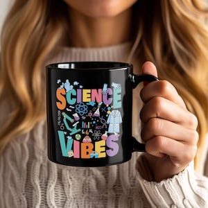 May include: Black ceramic mug with the words "SCIENCE VIBES" in colourful letters. The mug features science-themed illustrations, including a microscope, atoms, and lab coats. The mug is held by a person.