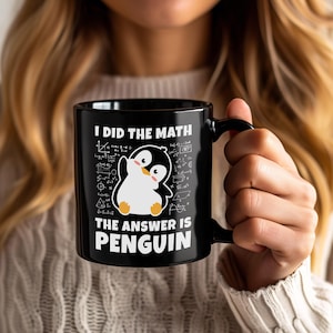 Op de afbeelding: Zwarte keramische mok met de tekst "I DID THE MATH THE ANSWER IS PENGUIN". De mok heeft een cartoon pinguïn illustratie en wiskundige vergelijkingen. De mok heeft een zwart handvat en wordt door een persoon vastgehouden.