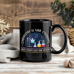 Puede incluir: Taza de cerámica negra con asa curva. La taza presenta el texto "CHEMISTRY IS LIKE COOKING" y "JUST DON'T LICK THE SPOON" con un gráfico de equipos de laboratorio. El diseño incluye vasos de precipitados, matraces y un tubo de ensayo.