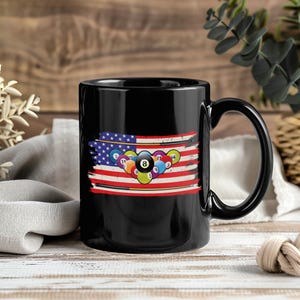May include: Black coffee mug featuring a distressed American flag design with a collection of billiard balls. The mug has a large handle and is set against a blurred background of wood and greenery.