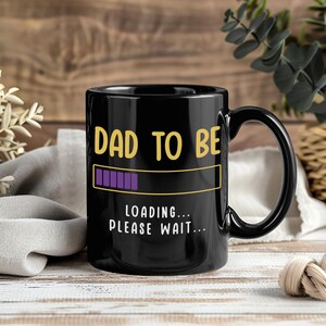 Peut inclure: Mug en céramique noire avec l'inscription dorée "DAD TO BE". En dessous, un graphique de barre de chargement avec des barres de progression violettes. Le mug porte également l'inscription "LOADING... PLEASE WAIT..." en blanc.