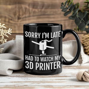 Könnte beinhalten: Schwarze Keramik-Tasse mit weißer 3D-Drucker-Grafik und dem Text "SORRY I'M LATE HAD TO WATCH MY 3D PRINTER". Die Tasse hat einen schwarzen Henkel und steht auf einer Holzoberfläche.