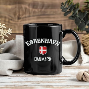 May include: Black coffee mug with the text "KØBENHAVN" above a red and white Danish flag emblem, and the word "DANMARK" below. The mug has a curved handle and is set against a wooden background.