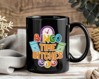 May include: Black ceramic mug with the text "BINGO TIME BITCHES" in vibrant colours. The design includes a clock, bingo cards, and bingo balls numbered 14, 7, and 2. A fun gift for bingo players.
