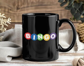 May include: Black ceramic mug with the word "BINGO" in colourful letters. The letters are in red, blue, green, yellow, and purple circles. The mug has a large handle and is set against a wooden background.