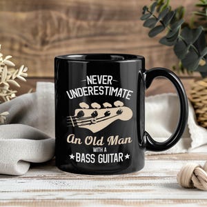 May include: Black ceramic mug with a curved handle. The mug features the text "NEVER UNDERESTIMATE An Old Man WITH A BASS GUITAR" in white and tan. A tan bass guitar headstock graphic is also present.