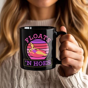 May include: Black ceramic mug with the text "FLOATS 'N HOES" in pink and purple. The mug features a graphic of a person floating on a pink inner tube in front of a sunset design.