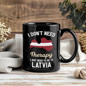 May include: Black ceramic mug with the text "I DON'T NEED Therapy I JUST NEED TO GO TO LATVIA". The mug features a graphic of the Latvian flag and country outline in red and white. The mug has a black handle and a glossy finish.