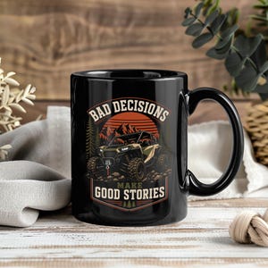 Könnte beinhalten: Ein schwarzer Keramikbecher mit einer grafischen Illustration eines Geländewagens vor Bergen und Bäumen. Der Text "BAD DECISIONS MAKE GOOD STORIES" ist in einer fetten, geschwungenen Schrift über und unter dem Fahrzeug abgebildet.