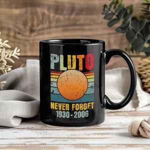 May include: Black ceramic mug with the word "PLUTO" in colorful retro lettering above a graphic of a planet. The mug also features the text "NEVER FORGET 1930-2006" in yellow. The mug has a black handle and is set against a wooden background.
