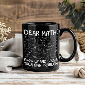 Peut inclure: Mug en céramique noire avec l'inscription blanche « DEAR MATH, GROW UP AND SOLVE YOUR OWN PROBLEM ». La tasse est recouverte d'équations et de symboles mathématiques. La tasse a une anse noire et est posée sur une surface en bois.