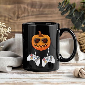 Op de afbeelding: Zwarte keramische mok met een Halloween-thema. Het ontwerp bevat een oranje pompoen met een zonnebril en een uitgesneden gezicht, die twee witte en grijze gamecontrollers vasthoudt. De mok heeft een zwart handvat.