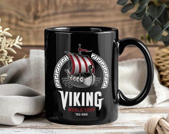 Puede incluir: Taza de cerámica negra con un diseño de barco vikingo, velas a rayas rojas y blancas y la palabra "VIKINGO" en blanco. Debajo, "WORLD TOUR 793-1066". La taza tiene un asa negra y un acabado brillante.