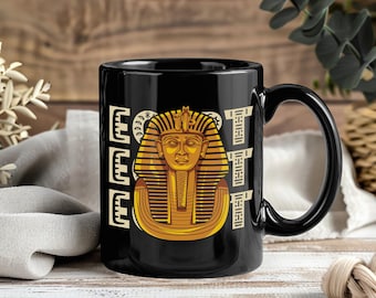 May include: Black ceramic mug featuring a gold and tan illustration of an Egyptian pharaoh's mask. The word "EGYPT" is printed vertically in a cream-colored font on the left and right sides of the mug. The mug has a black handle.