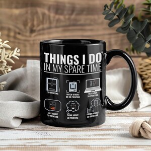 Könnte beinhalten: Schwarze Keramik-Tasse mit dem Text "THINGS I DO IN MY SPARE TIME" und Symbolen zum Thema 3D-Druck. Die Symbole zeigen einen 3D-Drucker, einen Laptop und Sprechblasen. Die Tasse hat einen schwarzen Henkel.