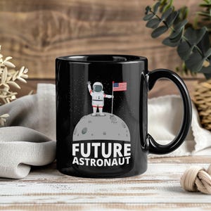 May include: Black ceramic mug with a cartoon astronaut on the moon holding an American flag. The mug has the words "FUTURE ASTRONAUT" in white. The mug has a black handle. The background has a starry design.