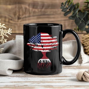 May include: Black ceramic mug featuring a stylized American flag design in the shape of a tree. The flag's stars and stripes are prominently displayed, with the roots of the tree in a red and white design. The mug has a black handle and a glossy finish.