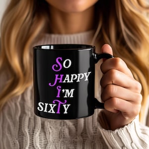 May include: Black ceramic mug with the text "So HAPPY I'm SIXTY" in white and purple lettering. The mug has a black handle and is held by a person wearing a white sweater.
