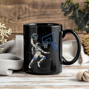 May include: A black ceramic mug featuring a graphic illustration of an astronaut in a white spacesuit dunking a planet Earth basketball into a blue basketball hoop against a dark starry space background with small white dots representing stars.