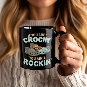 May include: Black coffee mug with the text "IF YOU AIN'T CROCIN' YOU AIN'T ROCKIN'" in light blue. The mug features illustrations of three different Crocs shoes. The mug is held by a person.