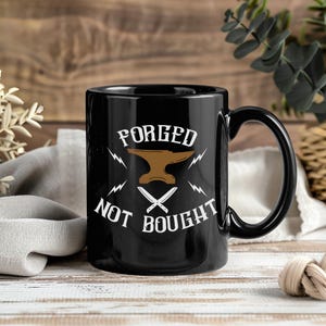 May include: Black ceramic mug with a handle, featuring the text "FORGED NOT BOUGHT" in white, along with a brown anvil and crossed knives design. The mug is set against a wooden background.