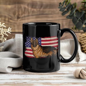 May include: Black ceramic mug with a German Shepherd dog design set against a distressed American flag. The dog is illustrated in brown and black. The mug has a large handle for easy gripping.
