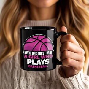 Può includere: Tazza da caffè nera con la scritta bianca "NEVER UNDERESTIMATE A GIRL WHO PLAYS BASKETBALL" e una grafica di pallacanestro rosa. La tazza è tenuta da una persona con lunghi capelli biondi.