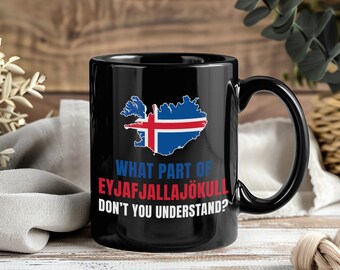May include: Black ceramic mug with the Iceland flag and the text "WHAT PART OF EYJAFJALLAJÖKULL DON'T YOU UNDERSTAND?". The mug has a black handle and is set against a wooden background.