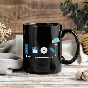 May include: Black ceramic mug featuring a white line with icons representing data storage: a server, floppy disk, CD, USB drive, and cloud. The mug has a black handle and a glossy finish. The design is a tech-themed graphic.
