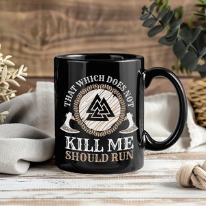 May include: Black ceramic mug with a large handle. The mug features the text "THAT WHICH DOES NOT KILL ME SHOULD RUN" around a central design of a triangle symbol and two axes. The design is in white and tan.